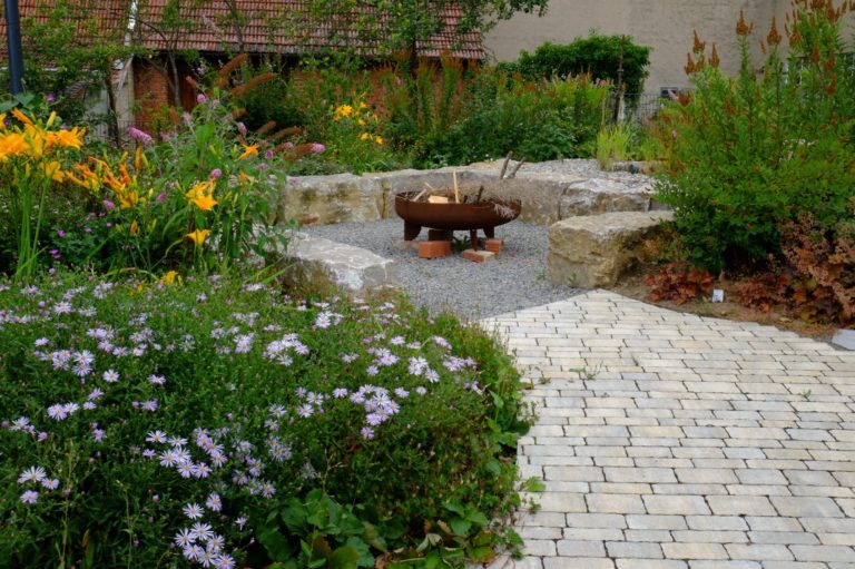 Gemütlicher Garten mit einer Feuerstelle, umgeben von bunten Blumen und Steinen.